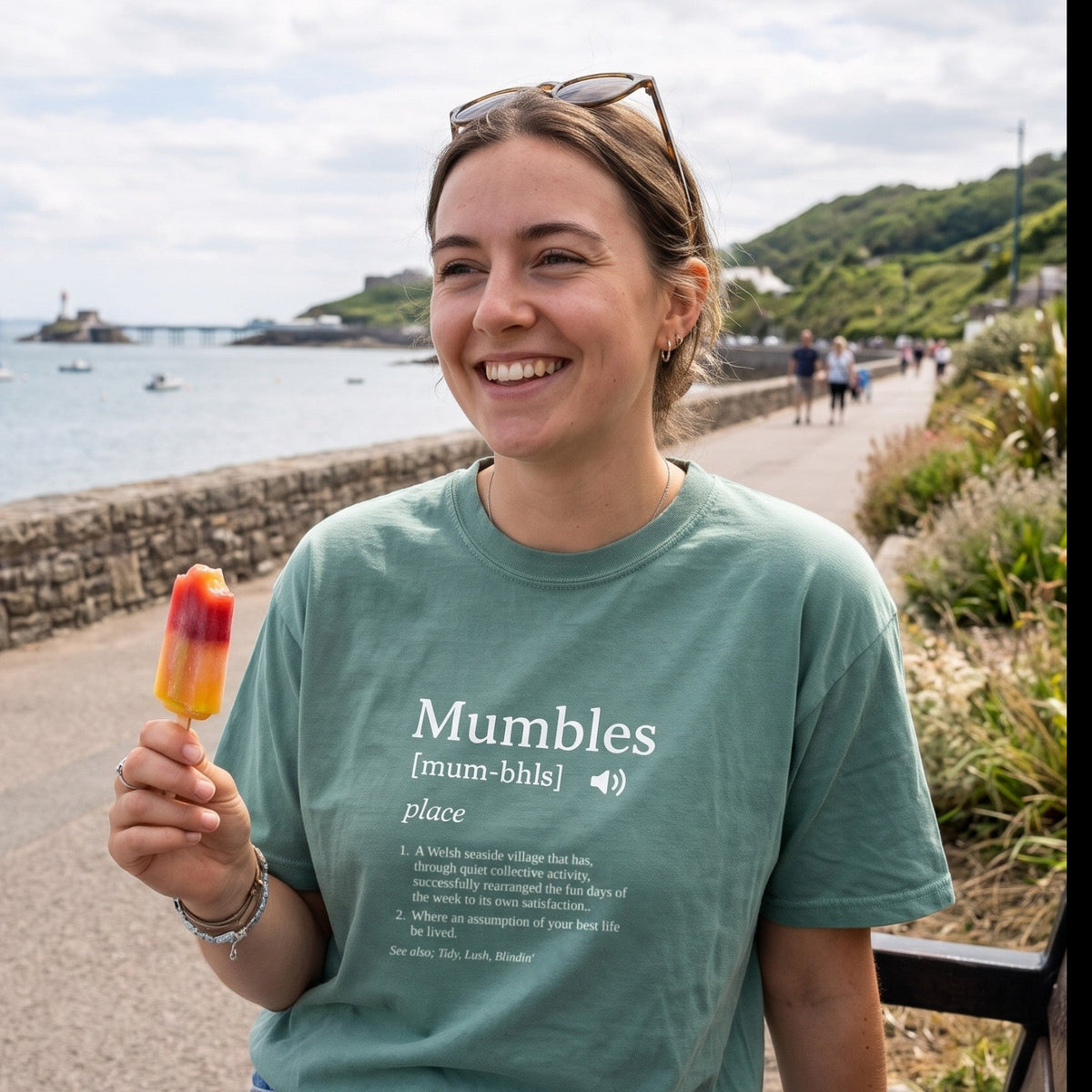 Woman on Mumbles promenade wearing the green Tidal Tings Mumbles definition T-shirt, with the lighthouse and pier behind